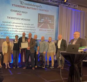 A group of people on stage hold a framed certificate with a congratulatory message to Kentucky Christian University displayed on a screen in the background.